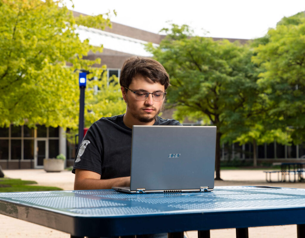 A person sits at a blue table outdoors, working on a laptop surrounded by trees and a building in the background.