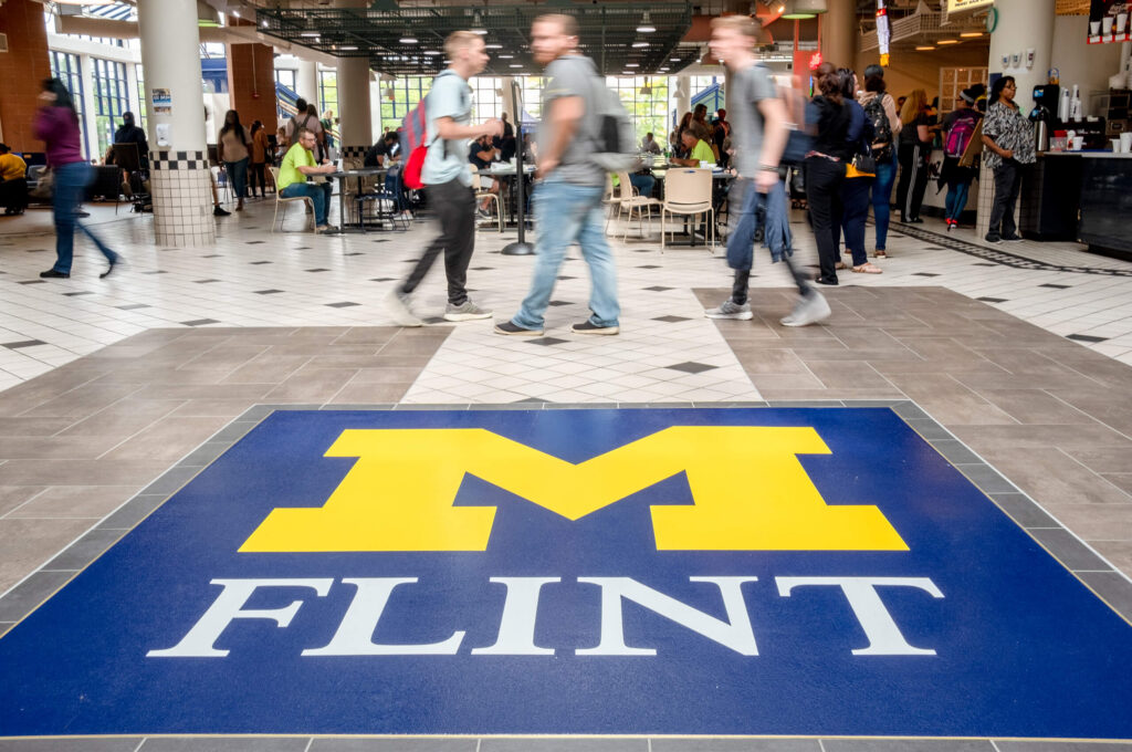A busy indoor space with people walking, featuring a large blue and yellow logo on the floor that reads "M FLINT."