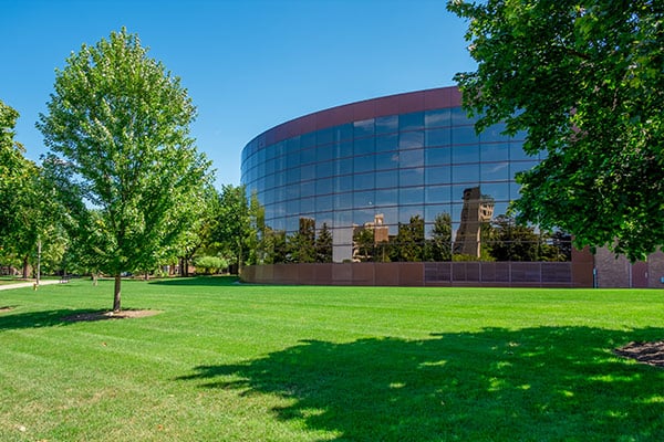 Modern curved glass building reflecting trees and a clear blue sky, surrounded by green grass and leafy trees.