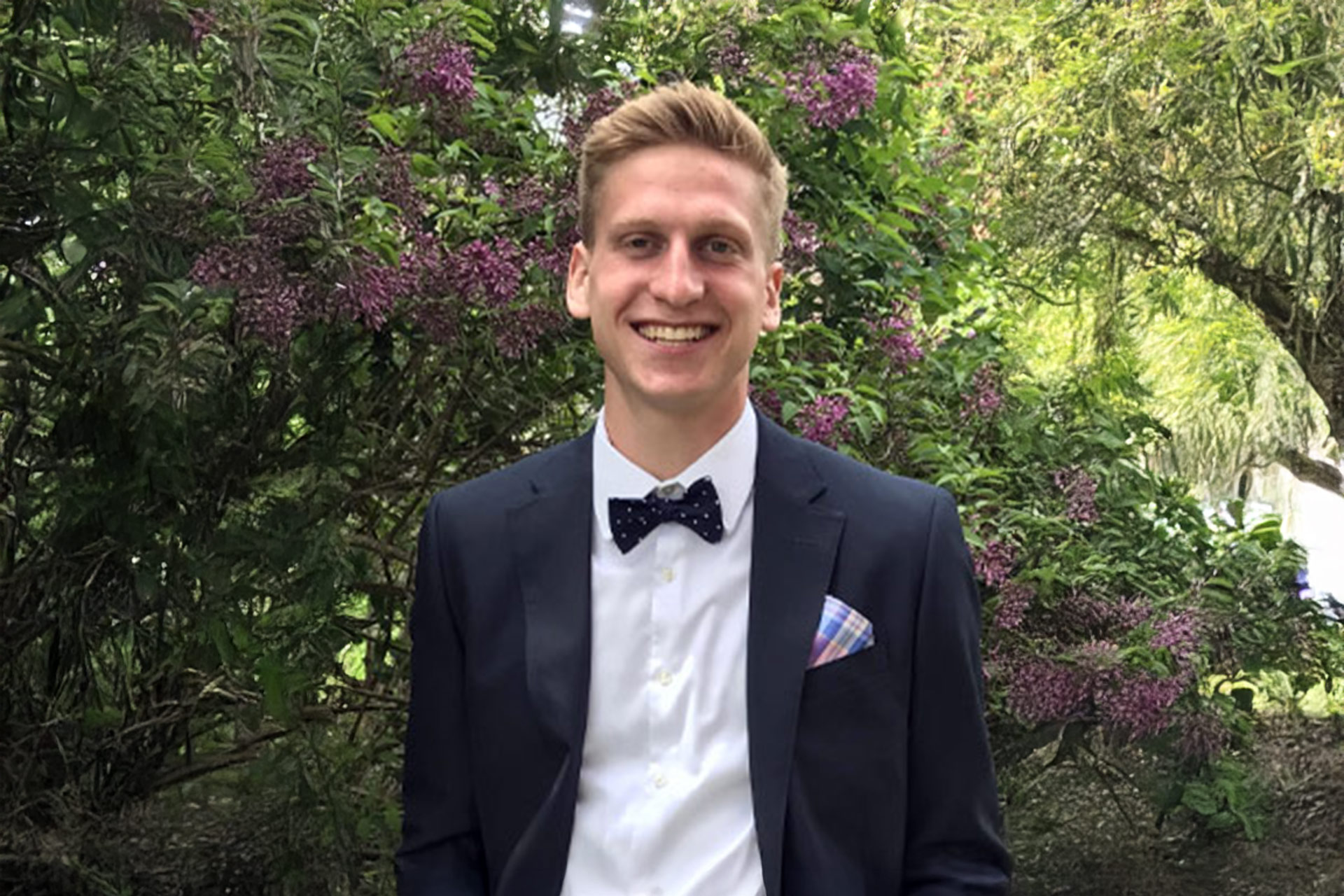 Larry Popyk in a formal suit with a bow tie stands in front of flowering bushes in a garden.