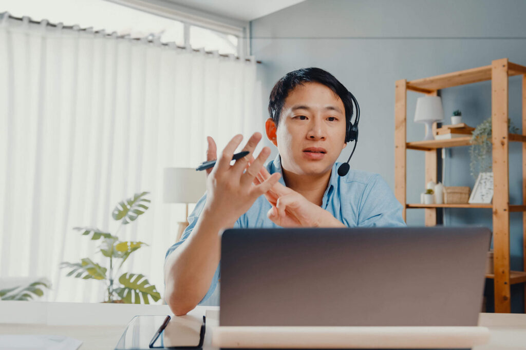 A person wearing a headset gestures while speaking during a video call, with a laptop in front of them.