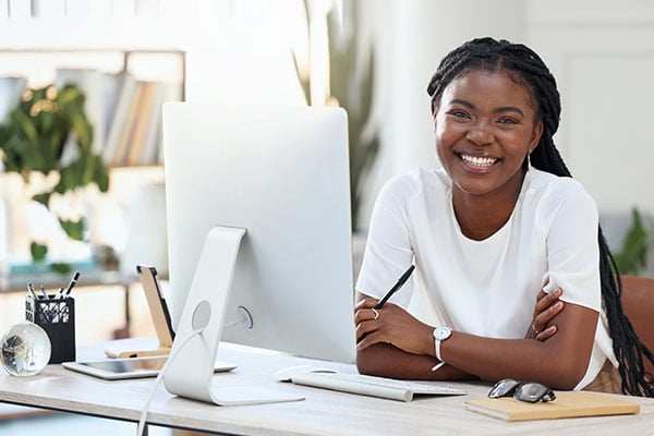A person sits at a desk with a computer, arms crossed, in a bright, modern workspace with plants in the background.
