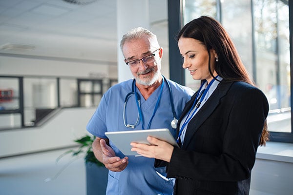 A person in scrubs and a stethoscope discusses information on a tablet with another person in a business suit.