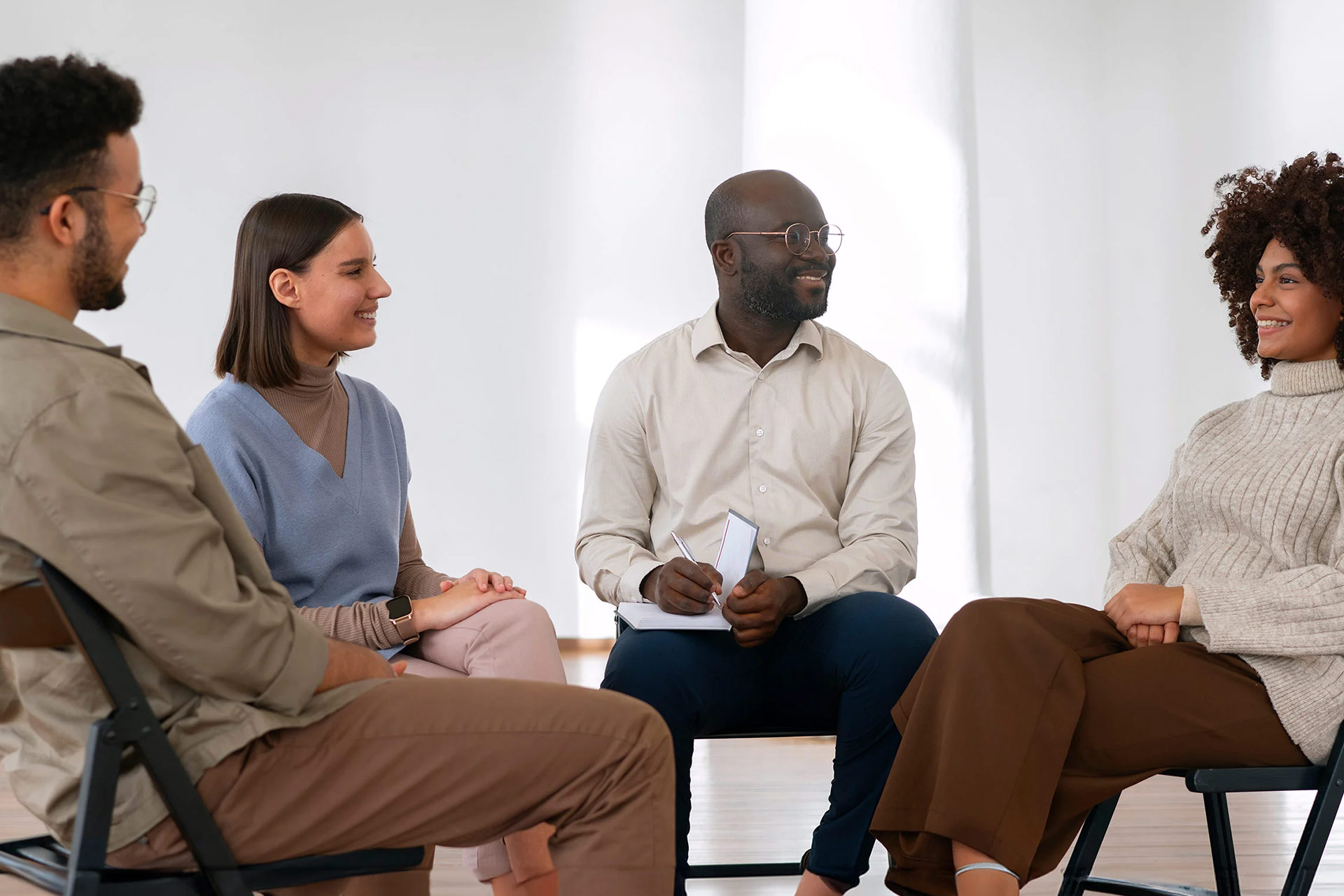 A group of four people sit in a circle, engaged in discussion, with one person taking notes.