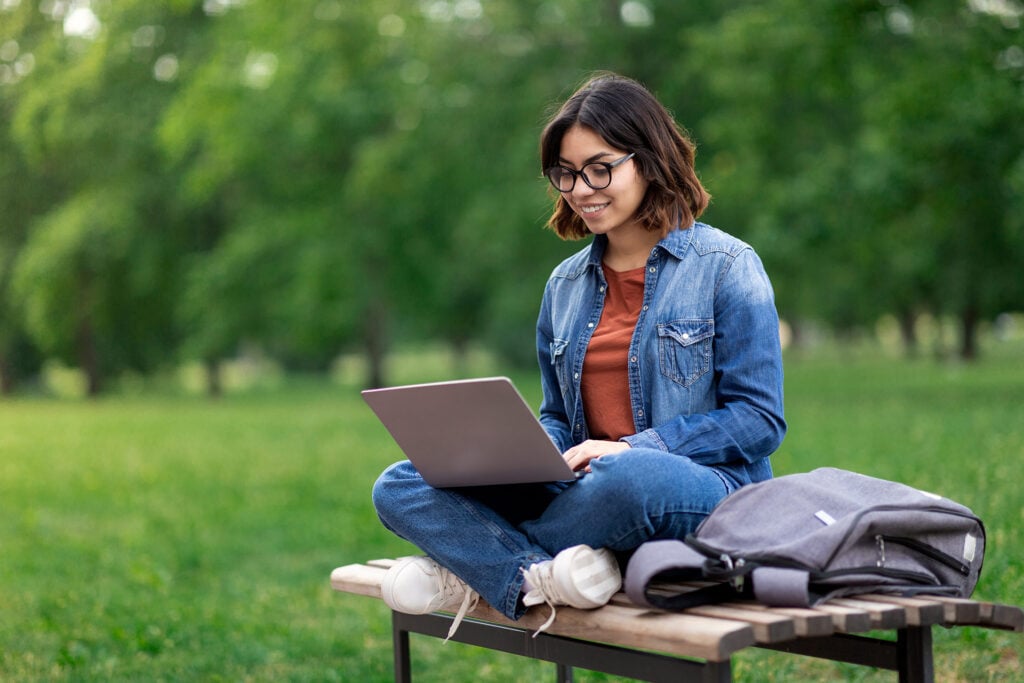 A person sits on a bench in a park, using a laptop with a backpack beside them.