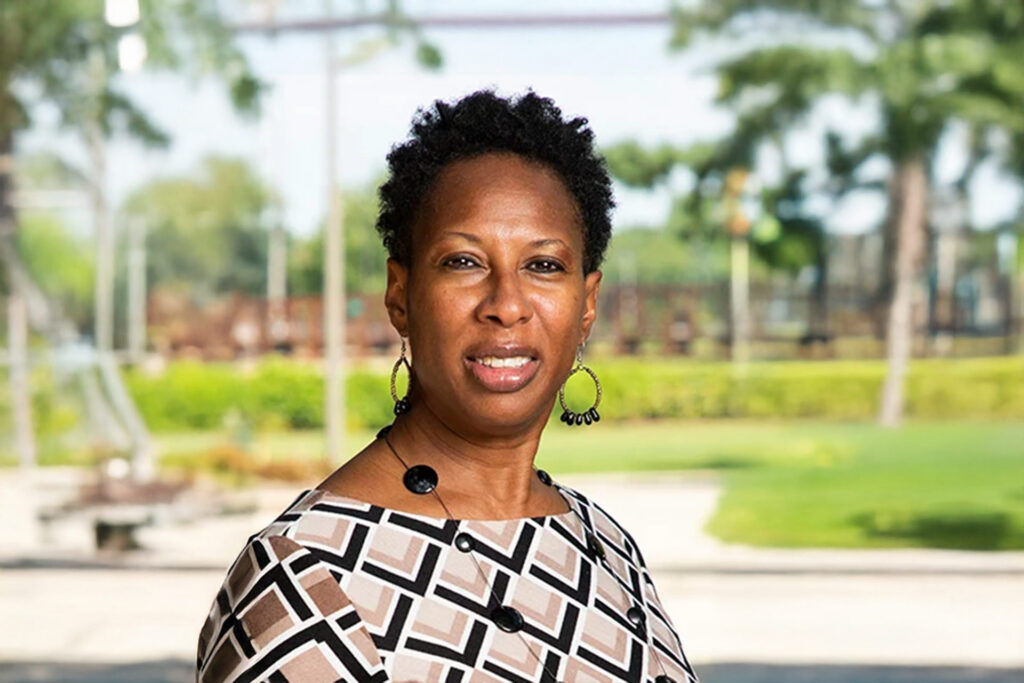Therasa Martin stands outdoors in a patterned top, with a blurred face, against a background of greenery and a playground.