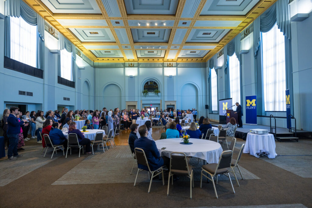 A large group of people is gathered in a spacious hall with tables, listening to a speaker at a podium, with banners for "M FLINT."