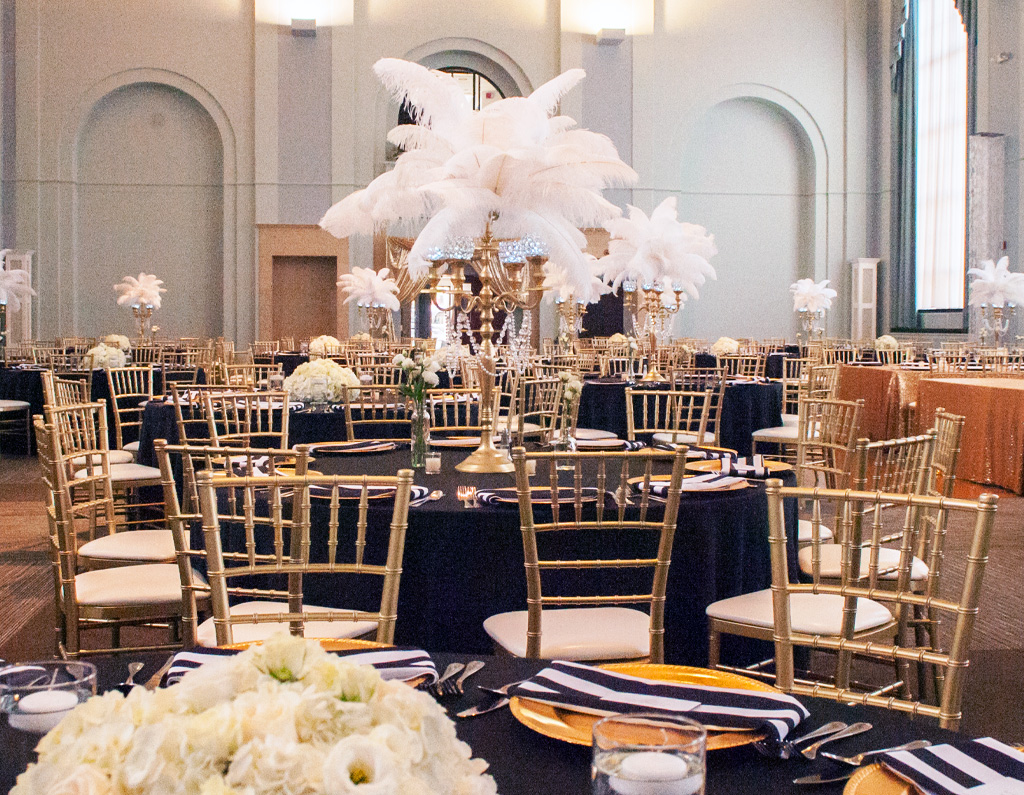 A banquet hall set for an event, featuring tables with black and gold decor, floral centerpieces, and tall feather arrangements.