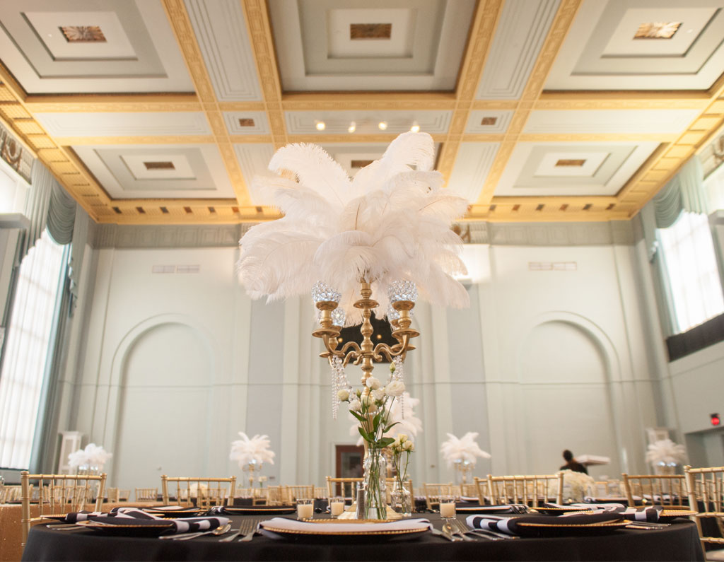 A beautifully decorated banquet hall featuring elegant tables with black tablecloths, gold chairs, and tall centerpieces with white feathers.