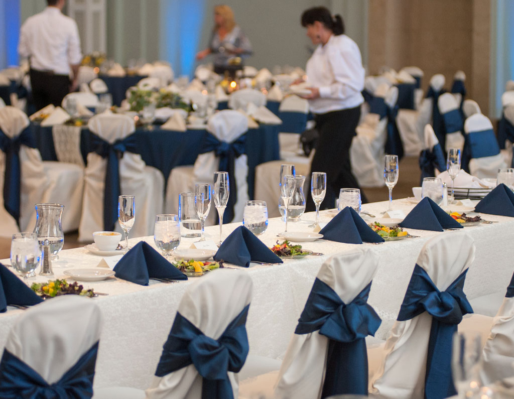 A banquet hall set for an event, featuring tables with blue and white decorations, glassware, and a server attending to guests.