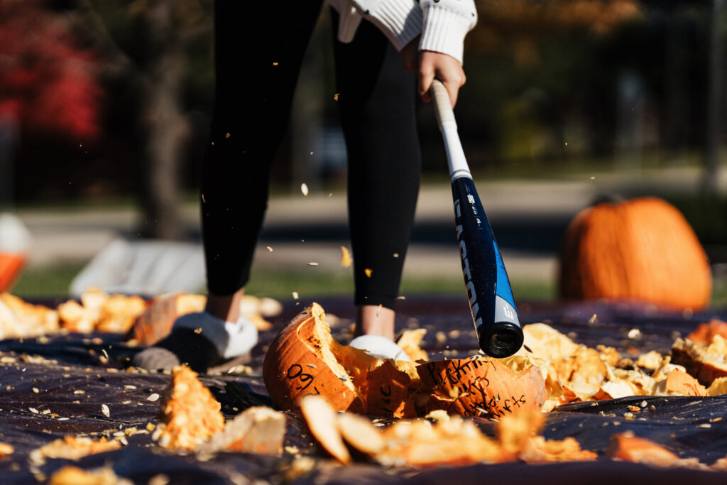 A person swings a bat to smash pumpkins on a black tarp, with pumpkin pieces and seeds flying around.