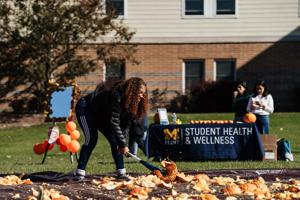 A person scoops pumpkin guts at an outdoor event, with a table displaying "Student Health & Wellness" in the background.