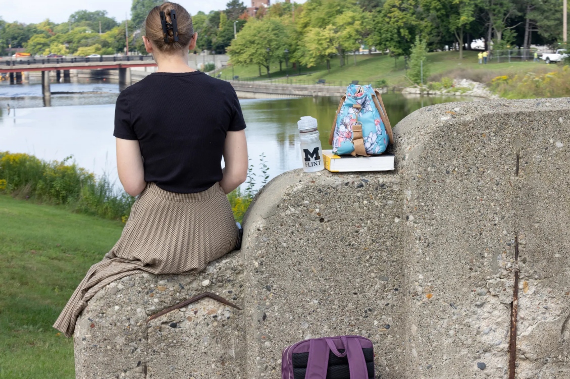 A person sits on a concrete barrier by a river, with a water bottle, a book, and a backpack beside them.