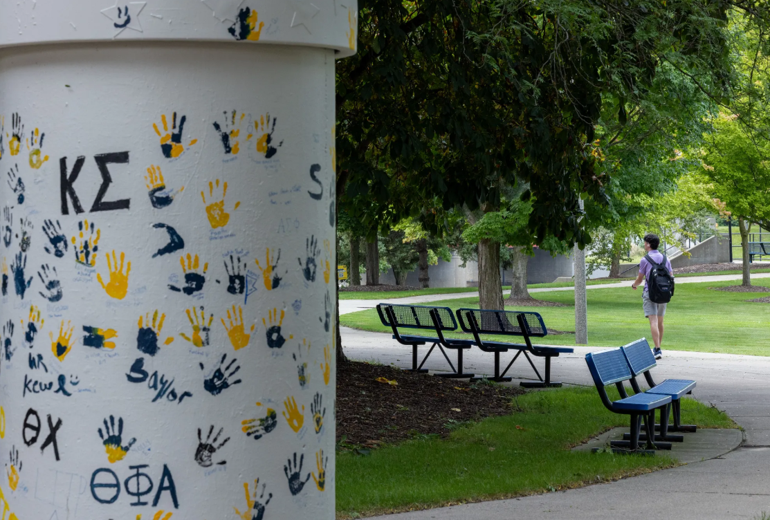 A column covered in handprints and letters stands in a park, with a person walking on a path nearby and benches in the foreground.