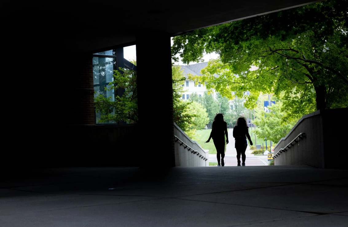 Two people walk together through a dark passageway, framed by greenery and light from the outside.