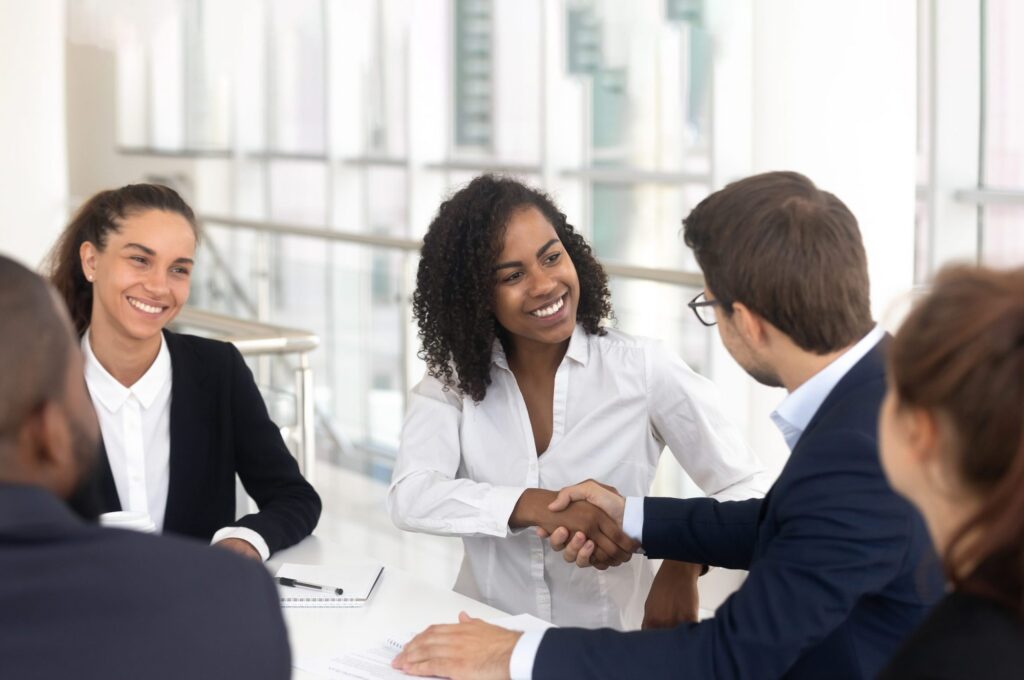 A group of people in a meeting, with two individuals shaking hands at a table.