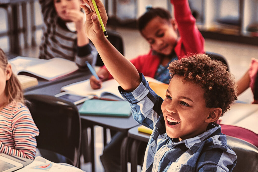 A group of children in a classroom, with one child raising their hand to answer a question.