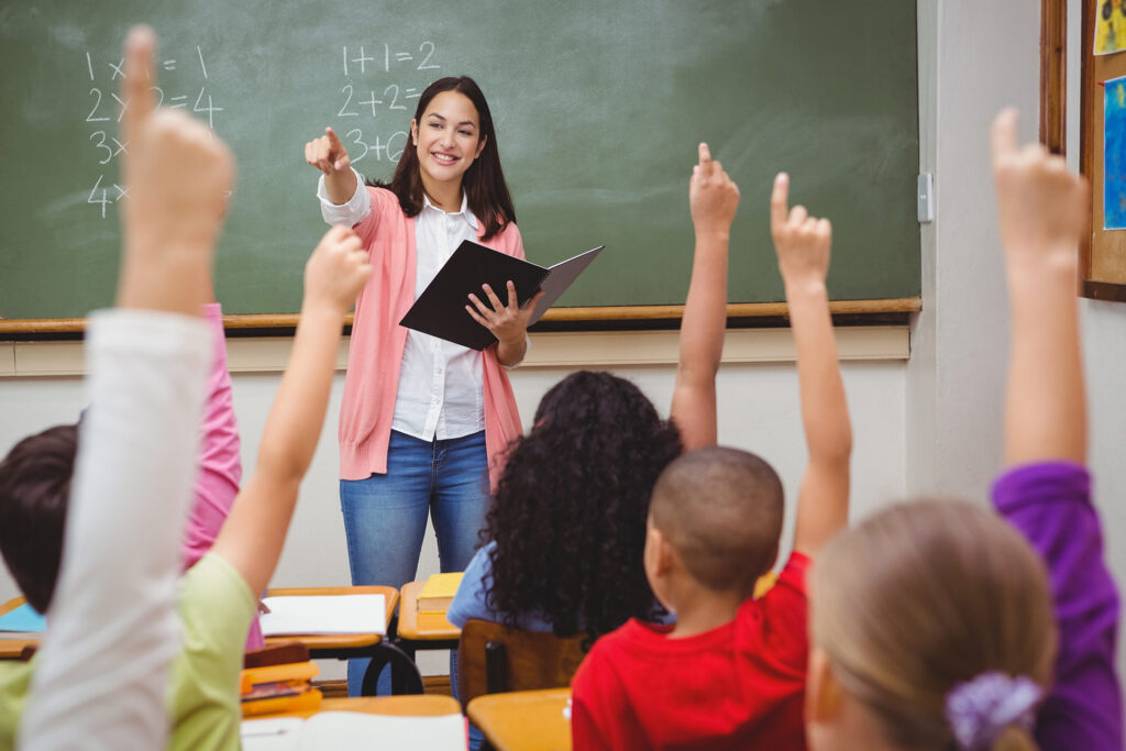 A teacher stands in front of a chalkboard, pointing while students raise their hands in a classroom setting.