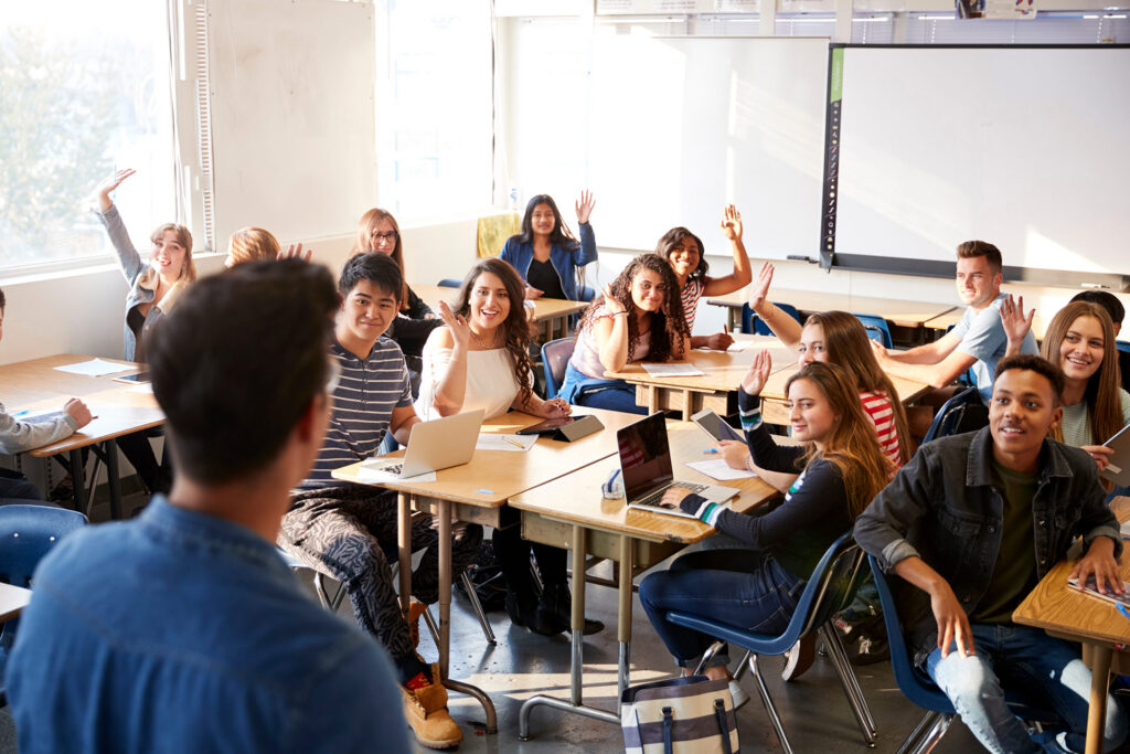 A group of students in a classroom raises their hands, engaging with a teacher who is facing them.