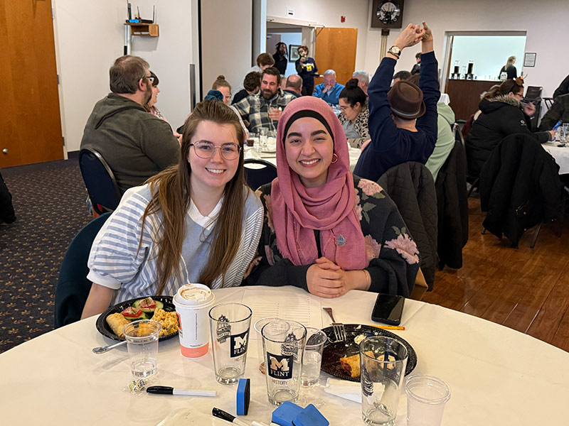 Two people smile at the camera while sitting at a table with food and drinks, surrounded by a busy dining room.