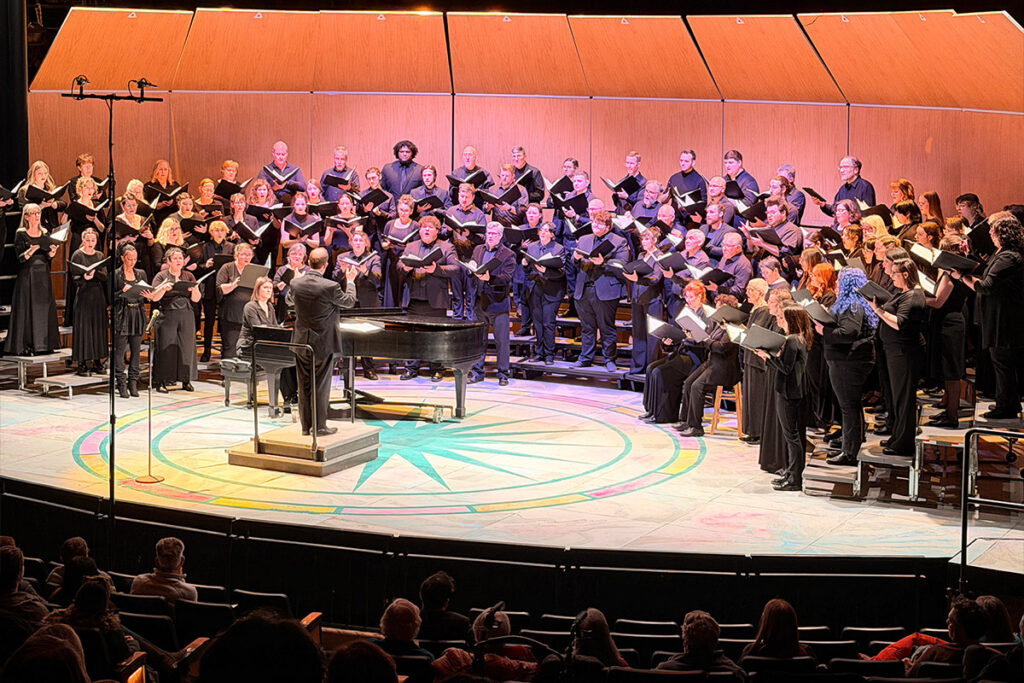 A large choir performs on stage, directed by a conductor at a piano, with an audience visible in the foreground.