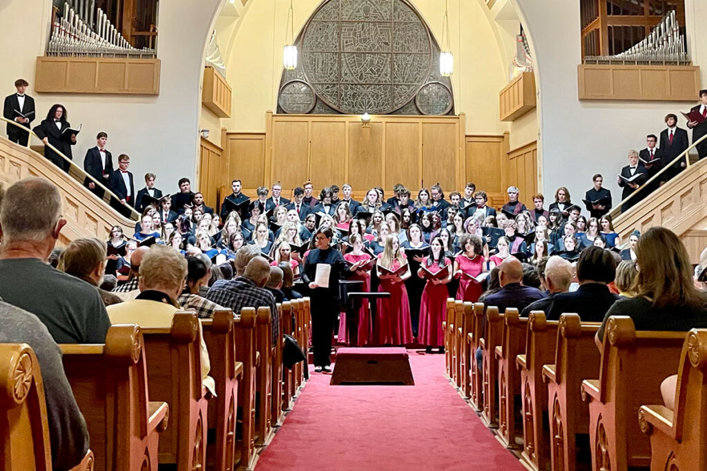 A large choir in formal attire performs on stage in a church, with an audience seated in front.