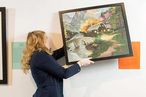 A person is hanging a framed painting of a landscape featuring a cabin, trees, and a stream on a wall in an exhibition space.