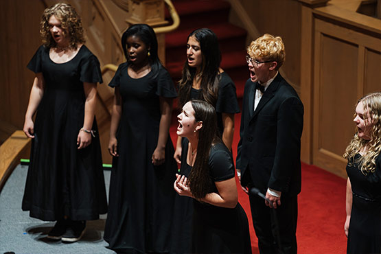 A group of six singers in black attire perform on stage, with one person passionately singing in the foreground.