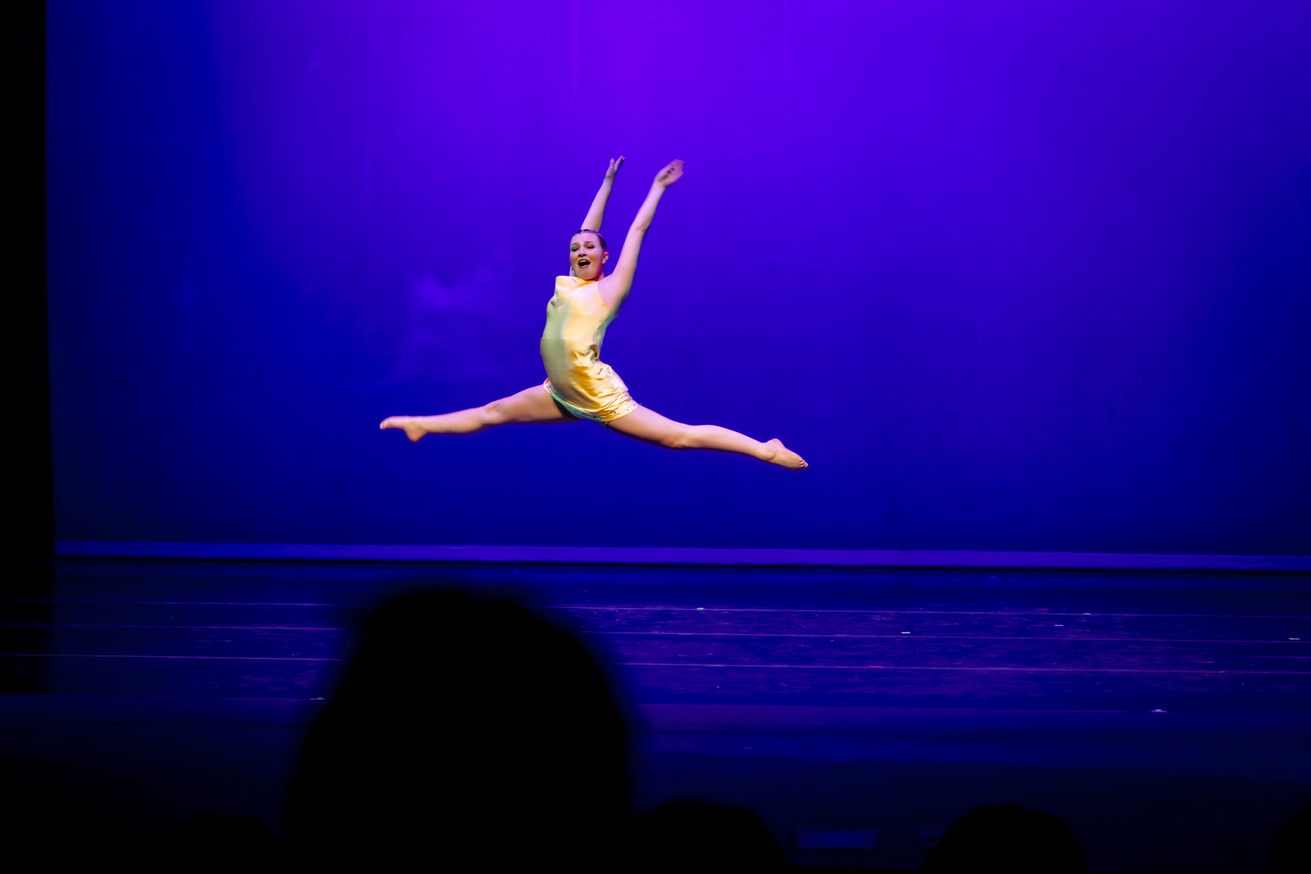 A dancer performs a leap on stage against a purple backdrop.