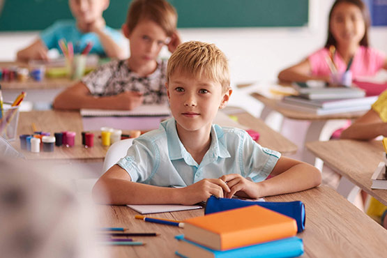 A child sits at a desk in a classroom, looking towards the front, with other students and school supplies visible in the background.