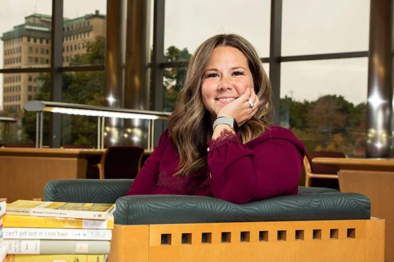 A person sitting in a library, resting their chin on their hand, with books stacked on a table in front of them.