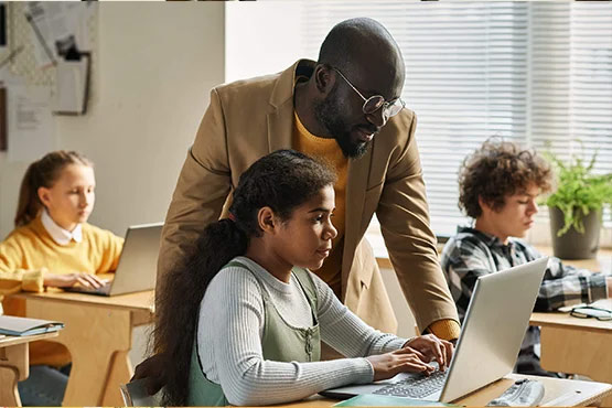 A person assists a student working on a laptop in a classroom, while two other students are focused on their devices.