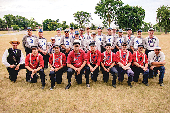 A group of twenty-two people in vintage baseball uniforms poses on a grassy field, with trees in the background.