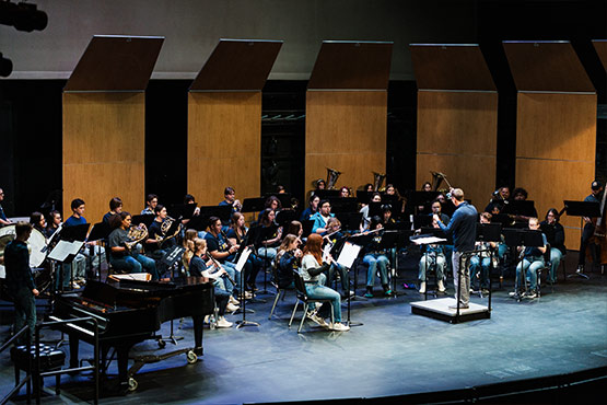 A conductor leads a large ensemble of musicians on stage, with a piano visible in the foreground.