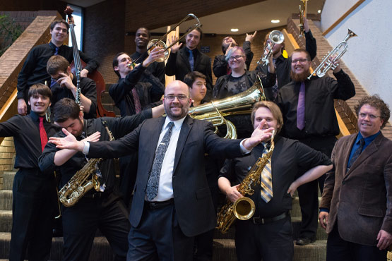 A group of musicians poses on stairs, holding various brass and woodwind instruments, with one person in front gesturing enthusiastically.