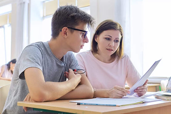 A person and a woman are sitting at a desk, discussing a paper while looking at each other in a classroom setting.