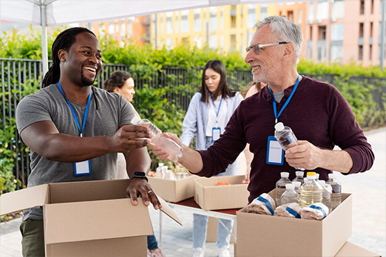 Two people are smiling and exchanging items while volunteering at an outdoor event, with boxes of supplies in front of them.
