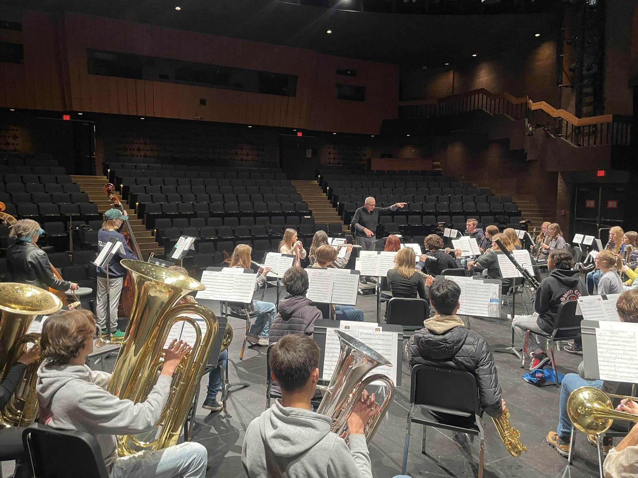 A conductor leads a group of musicians in a rehearsal on stage, with empty seats visible in the auditorium.