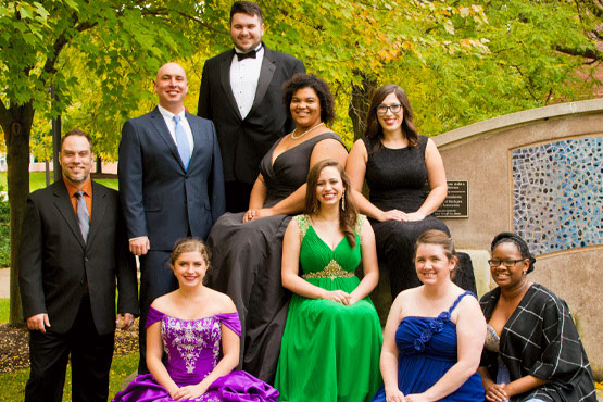 A group of nine people poses outdoors, dressed in formal attire, with trees and a decorative wall in the background.