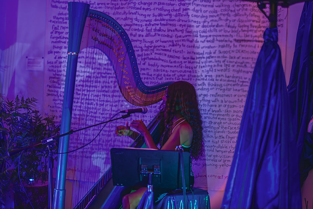 A person plays a harp while singing, with a wall covered in handwritten text and purple lighting in the background.