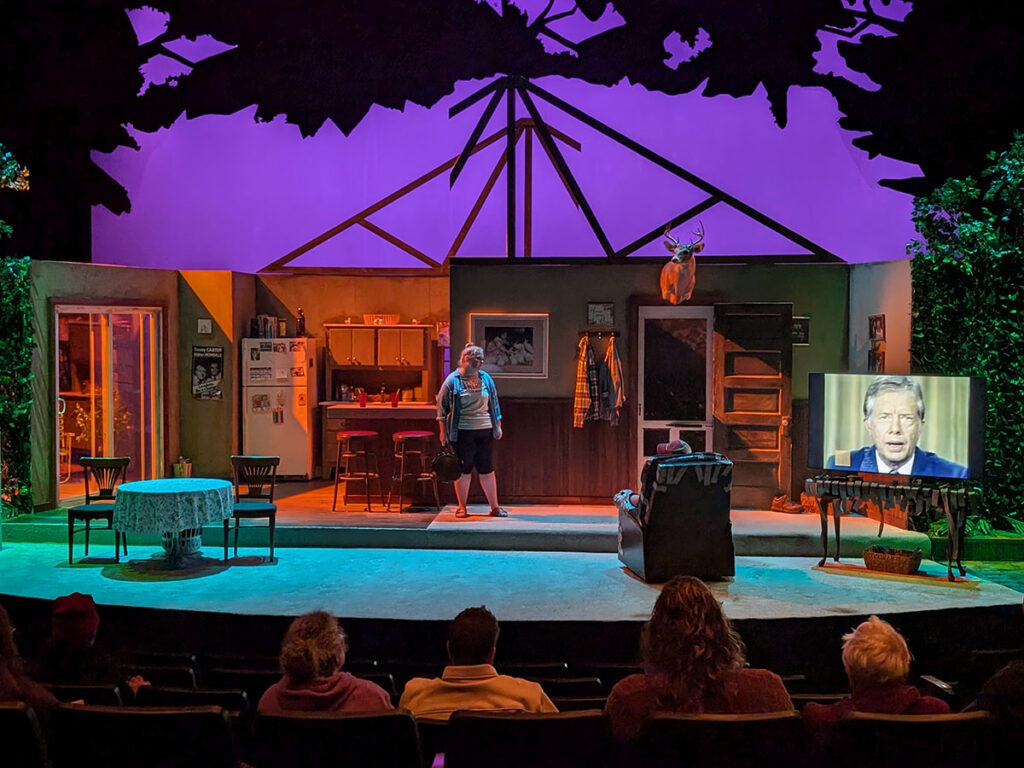 A stage set depicting a cozy cabin interior with a person standing, a television displaying a man speaking, and audience members visible.