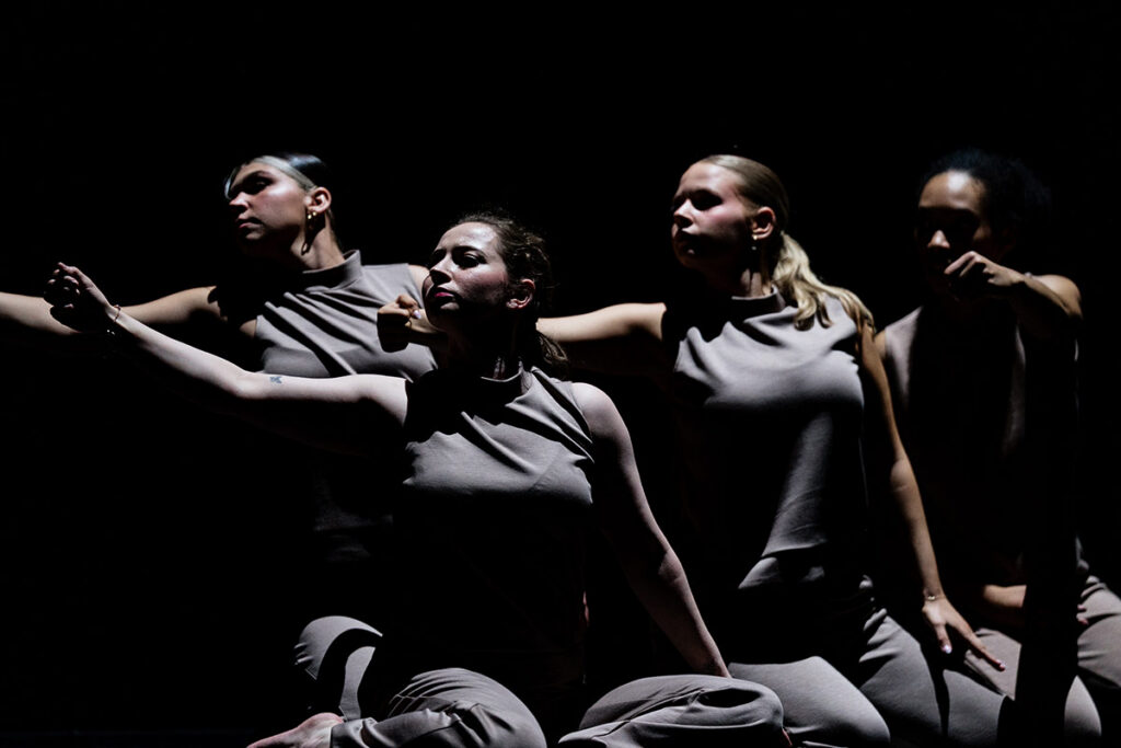 Four dancers in gray outfits perform expressively against a dark background, with arms extended and focused expressions.