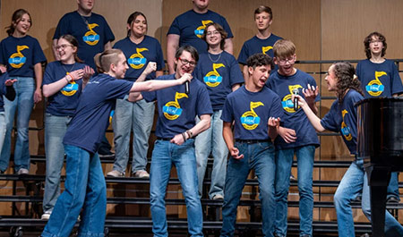 A group of people in matching blue shirts perform on stage, with some interacting playfully while others watch.