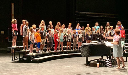 A group of children stands on risers in a theater, singing while a person conducts at a piano.