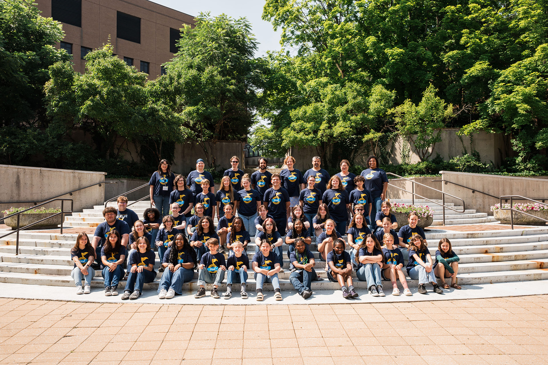 A large group of people in matching dark shirts poses on outdoor steps surrounded by greenery.