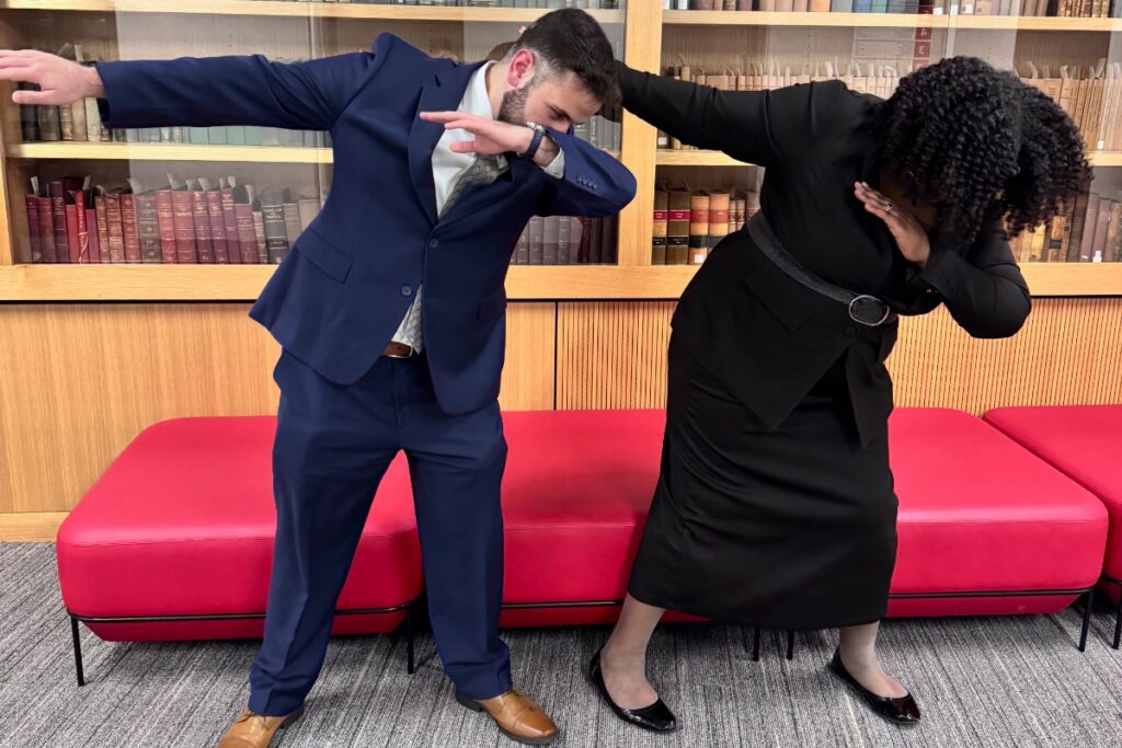 Two people are posing playfully, with one in a suit and the other in a black dress, both dabbing against a red couch backdrop.