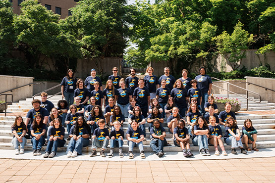 A large group of people, approximately 50, poses together on outdoor steps surrounded by greenery, all wearing matching t-shirts.