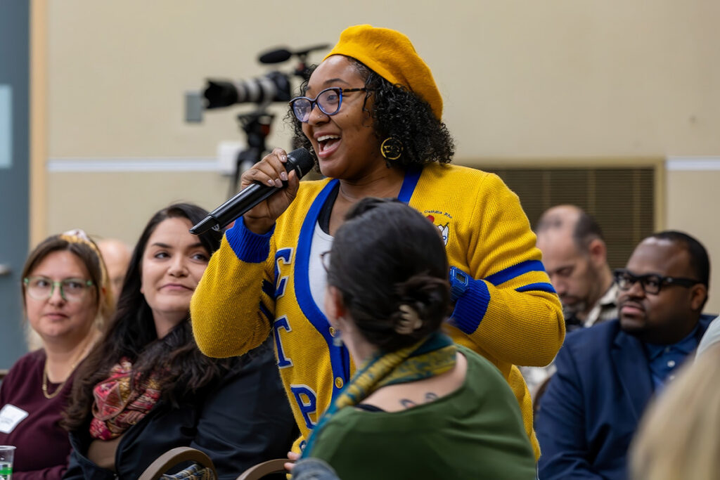 A person in a yellow beret and blue cardigan speaks into a microphone while seated among an audience.