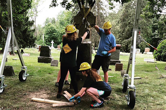 A group of four people, wearing hard hats, work together to stabilize a gravestone using equipment in a cemetery.