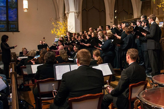 A choir performs in a church, with a conductor leading from the front and musicians seated in the foreground.